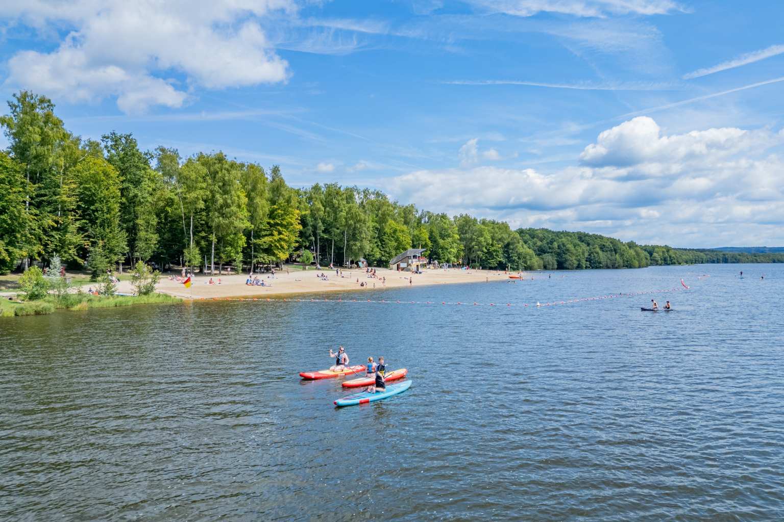 3 people in canoes paddling past the lake beach and forest next to Lac des Vielles Forges campsite in the Ardennes