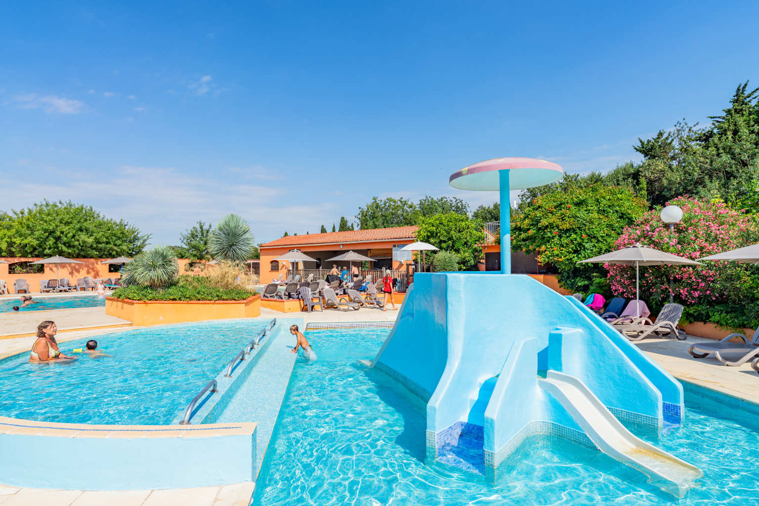 Parent and 2 small children in the semi circular pool next to paddling pool with small slides under mushroom feature at Ma Prairie campsite in the Roussillon