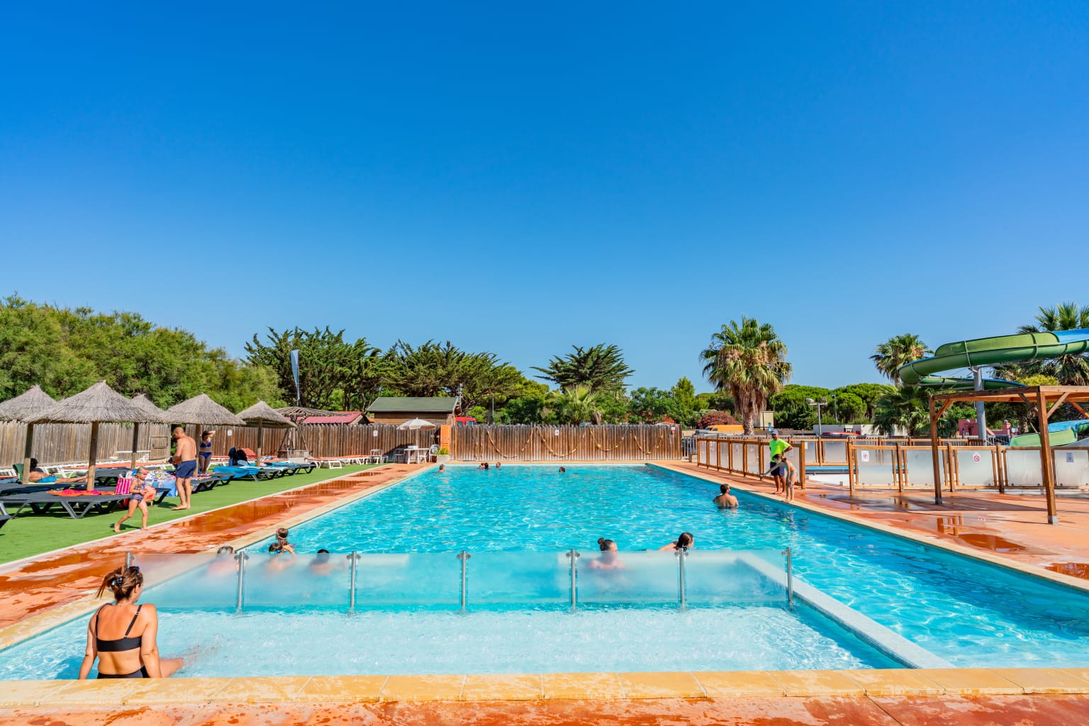 Lady sat on the edge of the shallow end of the rectangular outdoor pool, looking at the big waterslides on the right, at la Palmeraie campsite, Roussillon