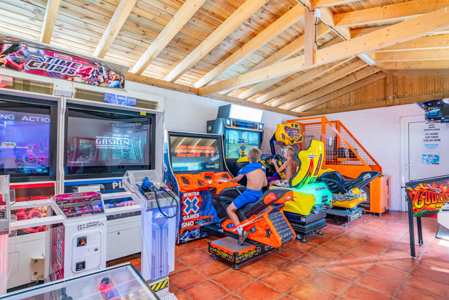 2 kids sat at games machines in the games barn, with wooden beamed ceiling, at le Soleil de la Mediterranee campsite, Roussillon