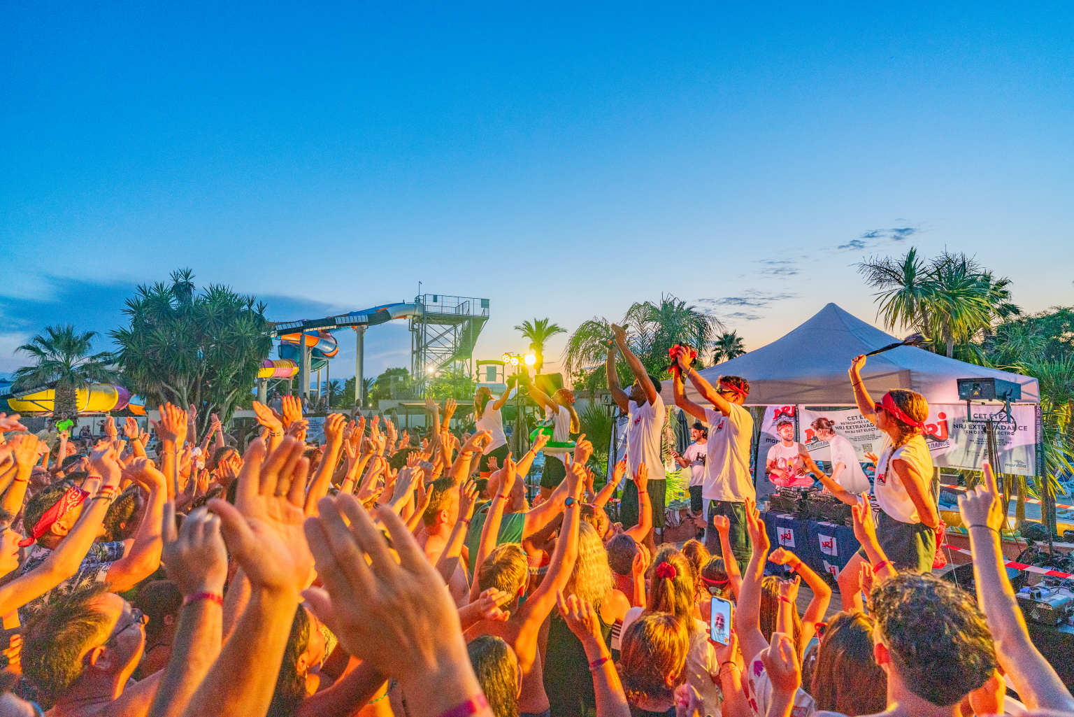 Crowd clapping hands to the beat at the outdoor disc with NRJ with view of waterslides at sunset at le Soleil de la Mediterranee campsite, Roussillon
