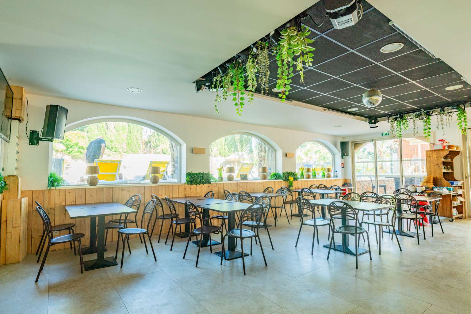 Tables and chairs inside the restaurant, next to arched windows, with artifical plants hanging from ceiling, at La Croix du Sud campsite in the Languedoc