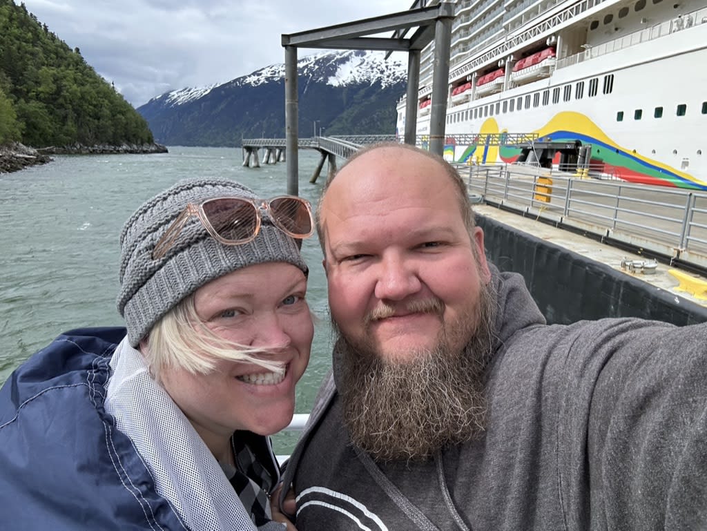 Two smiling individuals wearing casual clothing and a beanie near a dock with a large cruise ship in the background.