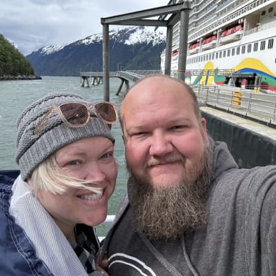 Two smiling individuals wearing casual clothing and a beanie near a dock with a large cruise ship in the background.