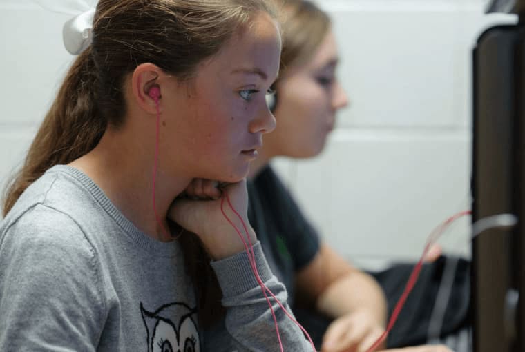 Two girls in class using headphones to listen to their desktop computers.