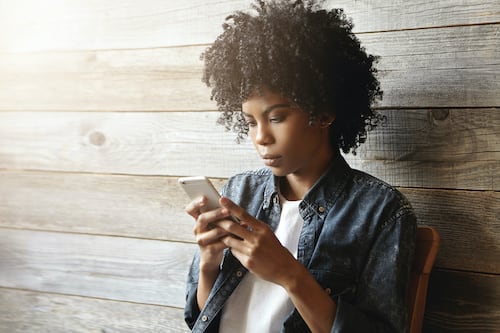 Beautiful young African hipster girl with curly hair wearing trendy clothing holding mobile phone, reading sms from her boyfriend, having serious and worried facial expression. People and technology Beautiful young African hipster girl with curly hair wearing trendy clothing holding mobile phone, reading sms from her boyfriend, having serious and worried facial expression. People and technology
