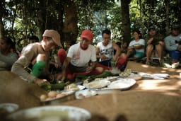 A group of men, women and children are sitting in the middle of the rainforest; they are seated around grassmat whch has large plates of fresh tropical fruit and breads.