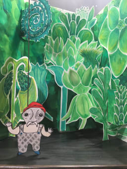 A small mustached paper puppet wearing a red hat and checked overalls stands in a world of green, a popup garden. 