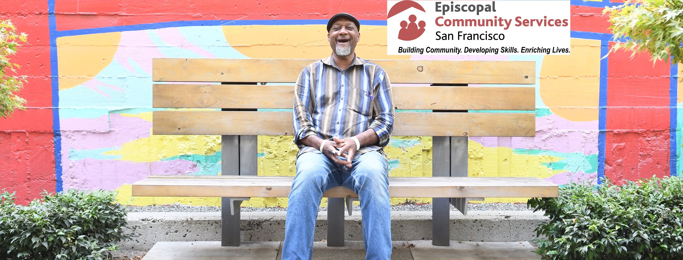 Image: Man in a striped shirt and jeans sits on a bench in front of a colorful mural.  His has a broad smile on his face. 