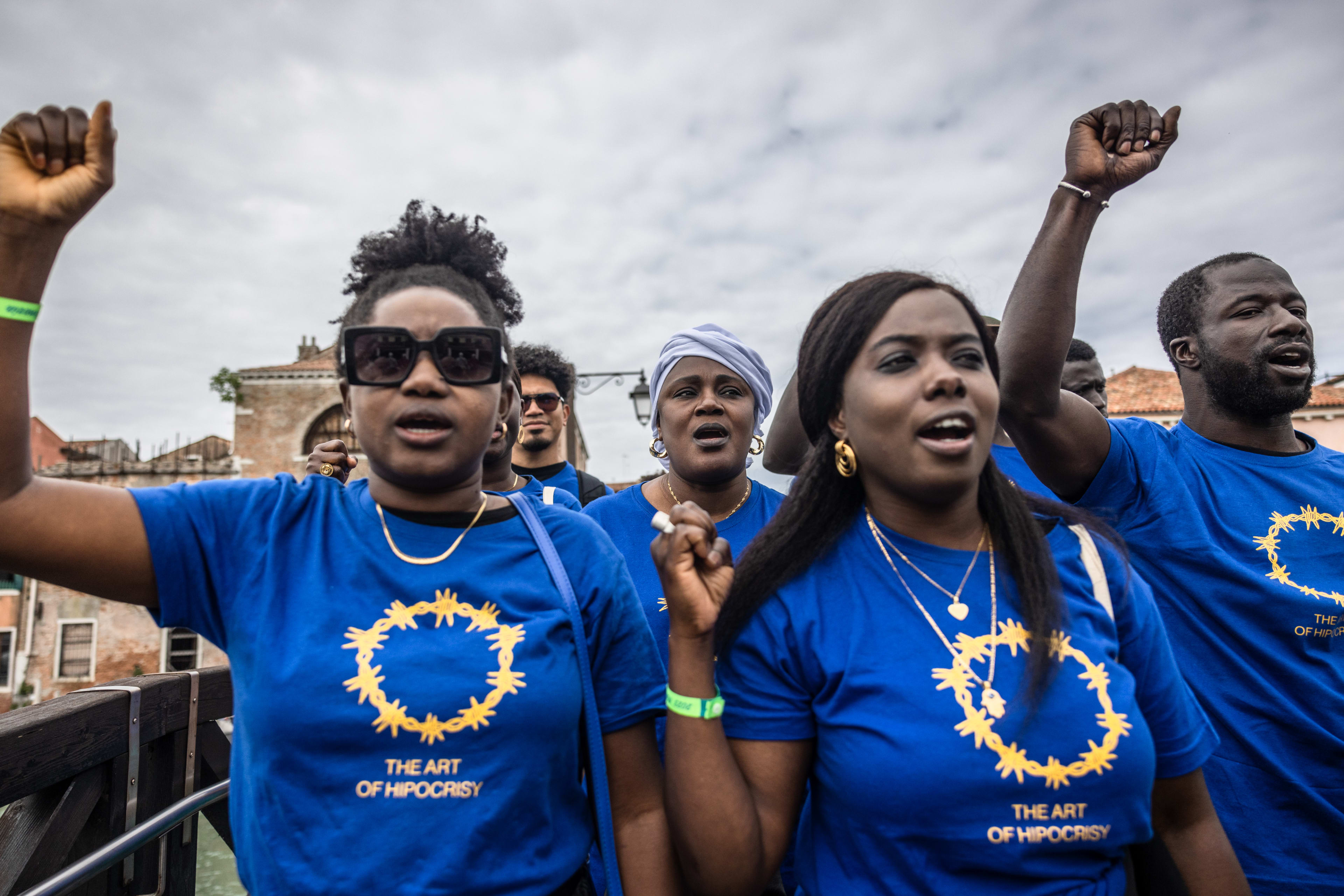 A group of Black migrants raise their fists in the air as they protest, wearing matching blue shirts that say "The Art of Hypocrisy" beneath yellow barbed wire circle, a reworking of the European Union symbol