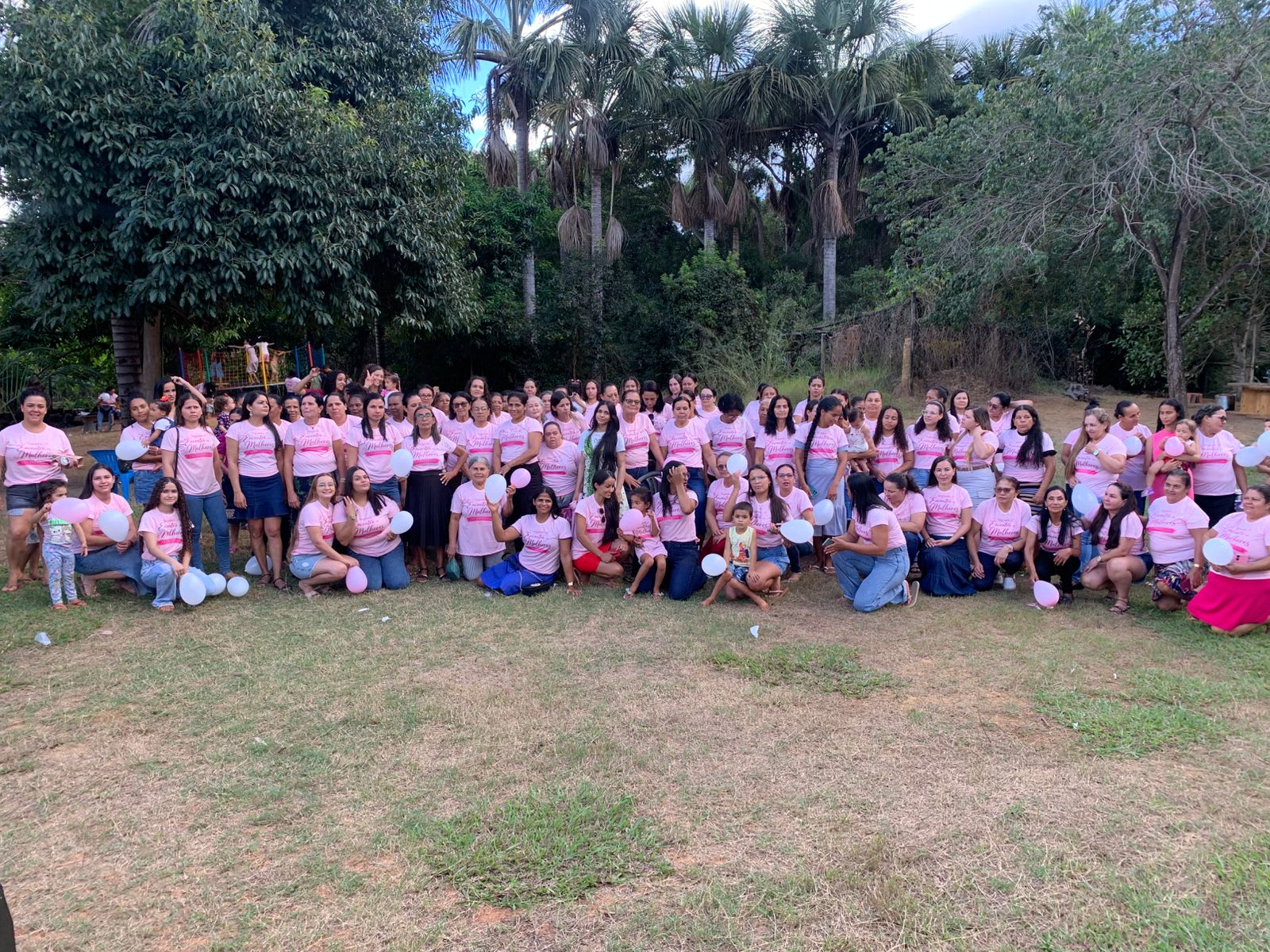 Women and children gathered outdoors for a group photo, many wearing pink shirts, with trees and palms in the background.