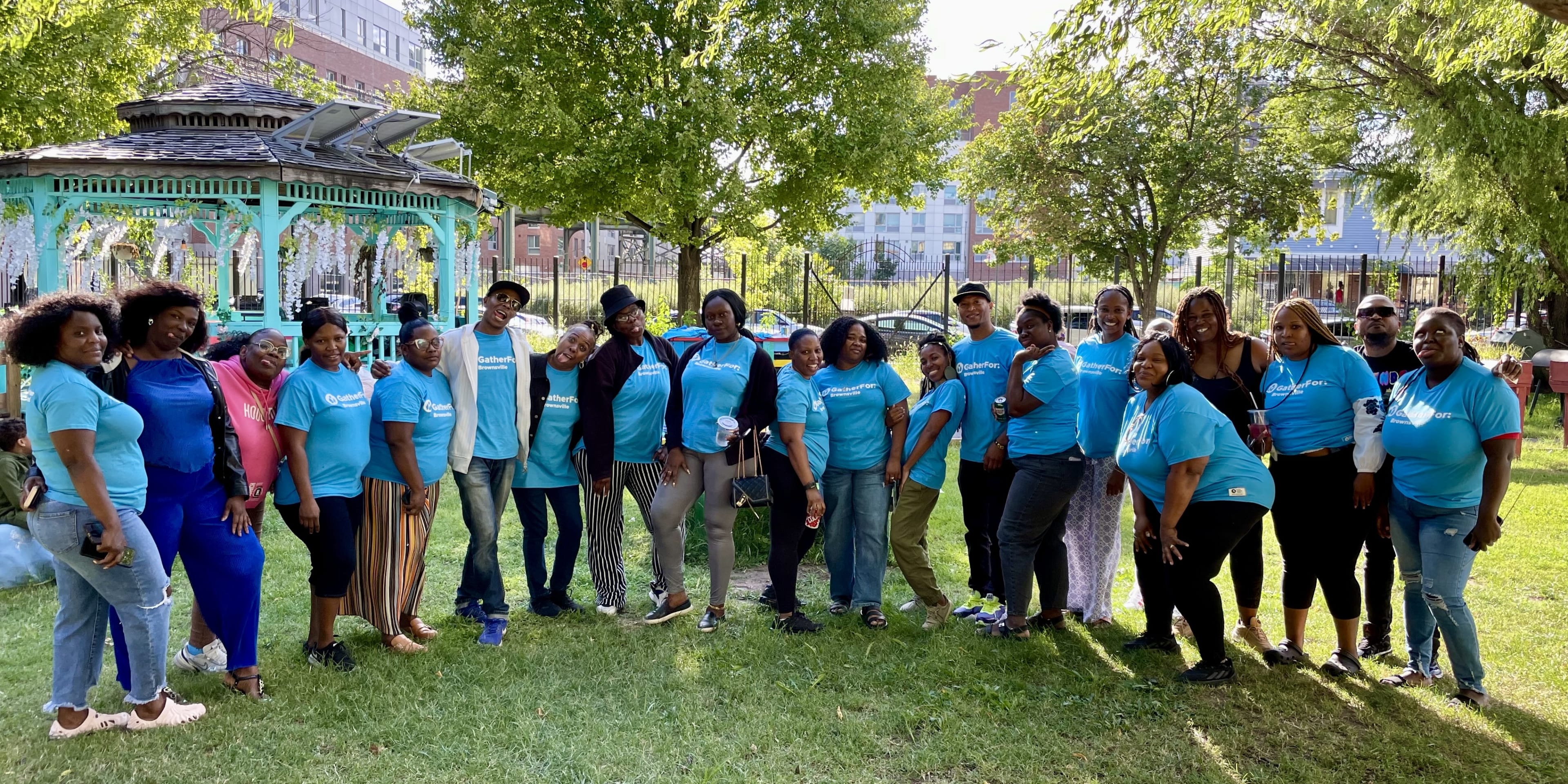 20 GatherFor participants wearing blue GatherFor t-shirts gather in a park under the shade of trees on a sunny day.
