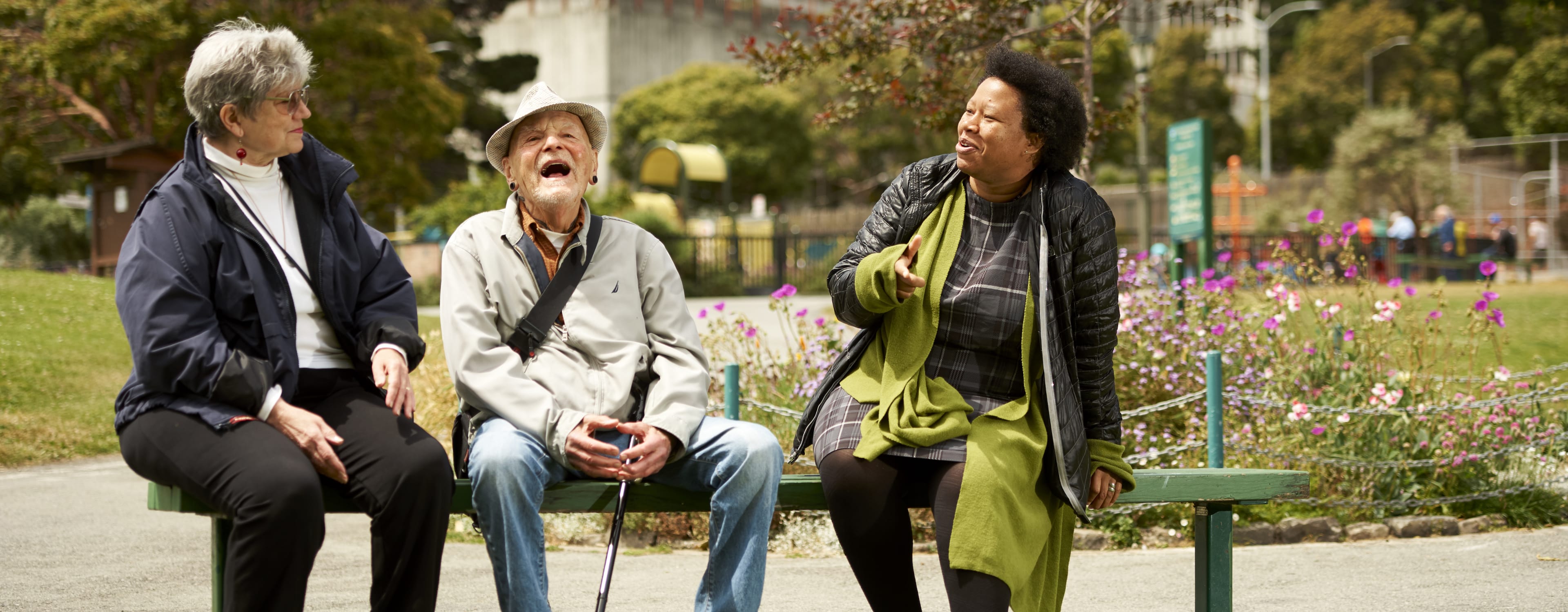 Three older adults, two women and one man, sit on a park bench. The man in the middle has his mouth open in a laugh or shout, while the two women look towards him, one smiling and one with an expressive, engaged face.