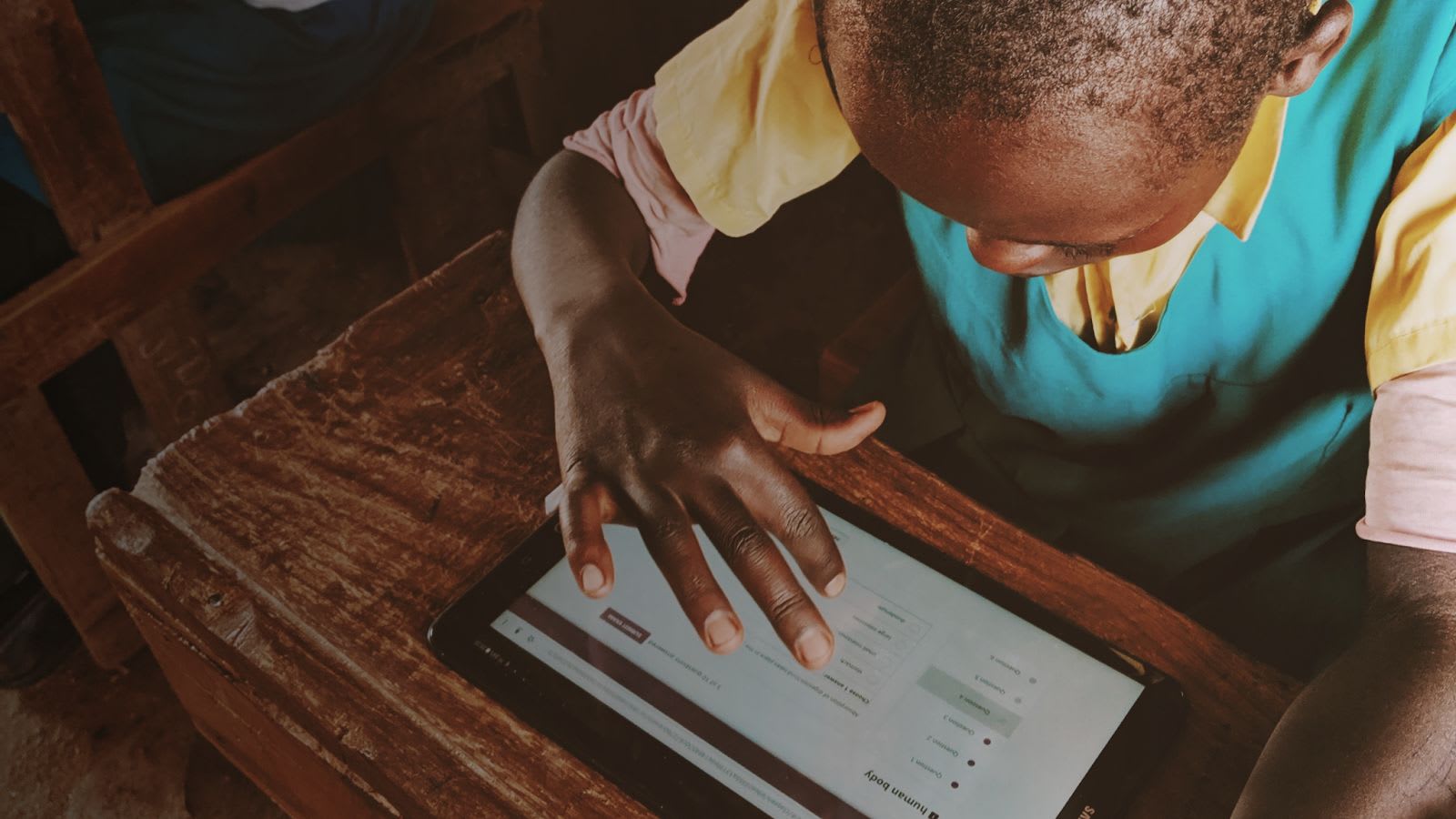 From above, we see the close-up of a learner sitting at his school desk, solving a math problem on a tablet. He’s in his school uniform, blue and yellow, the desk is a wooden desk.