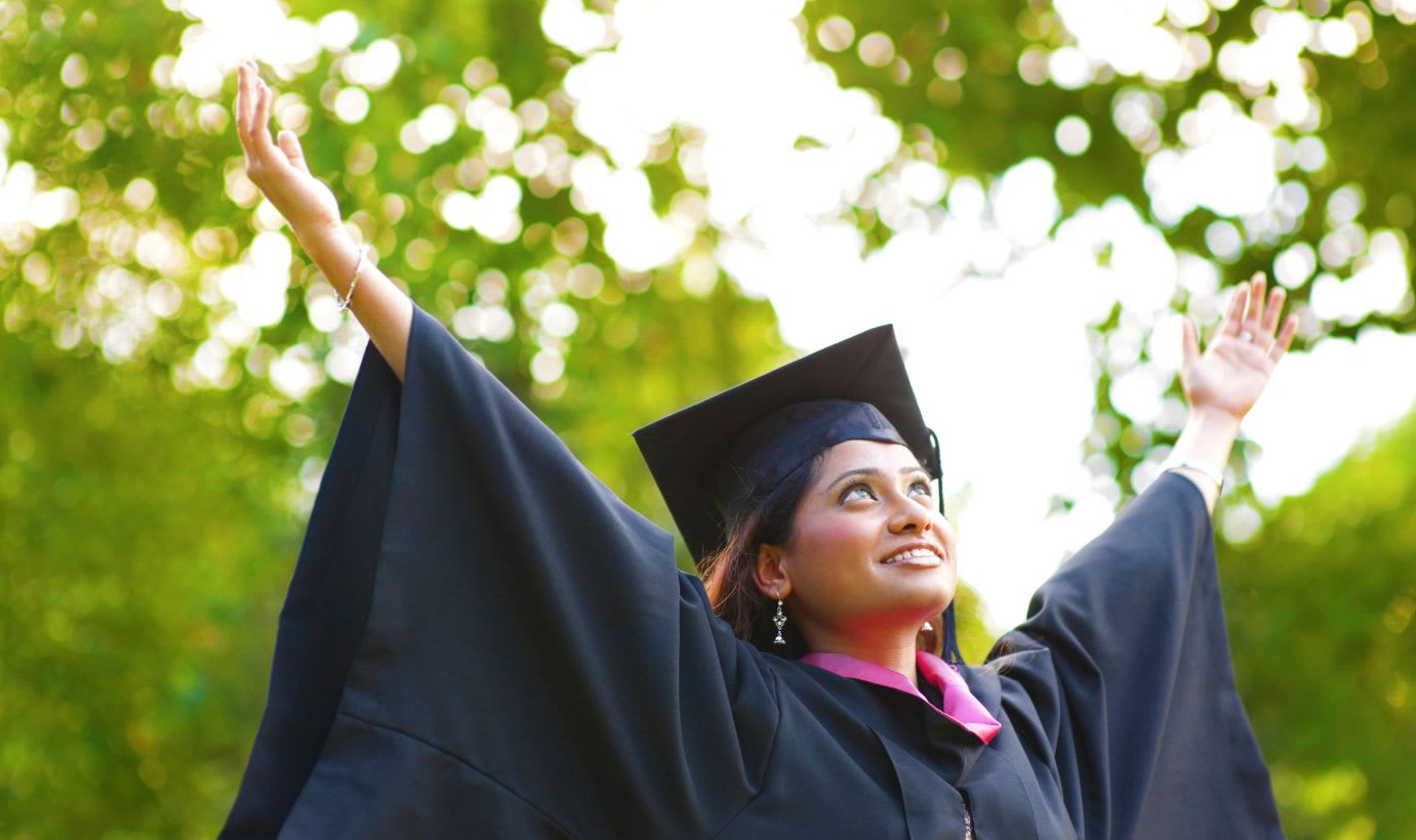 Young woman of Asian descent holds her hands outstretched to the sky wearing a university graduation cap and gown. Trees are visible in the background. 