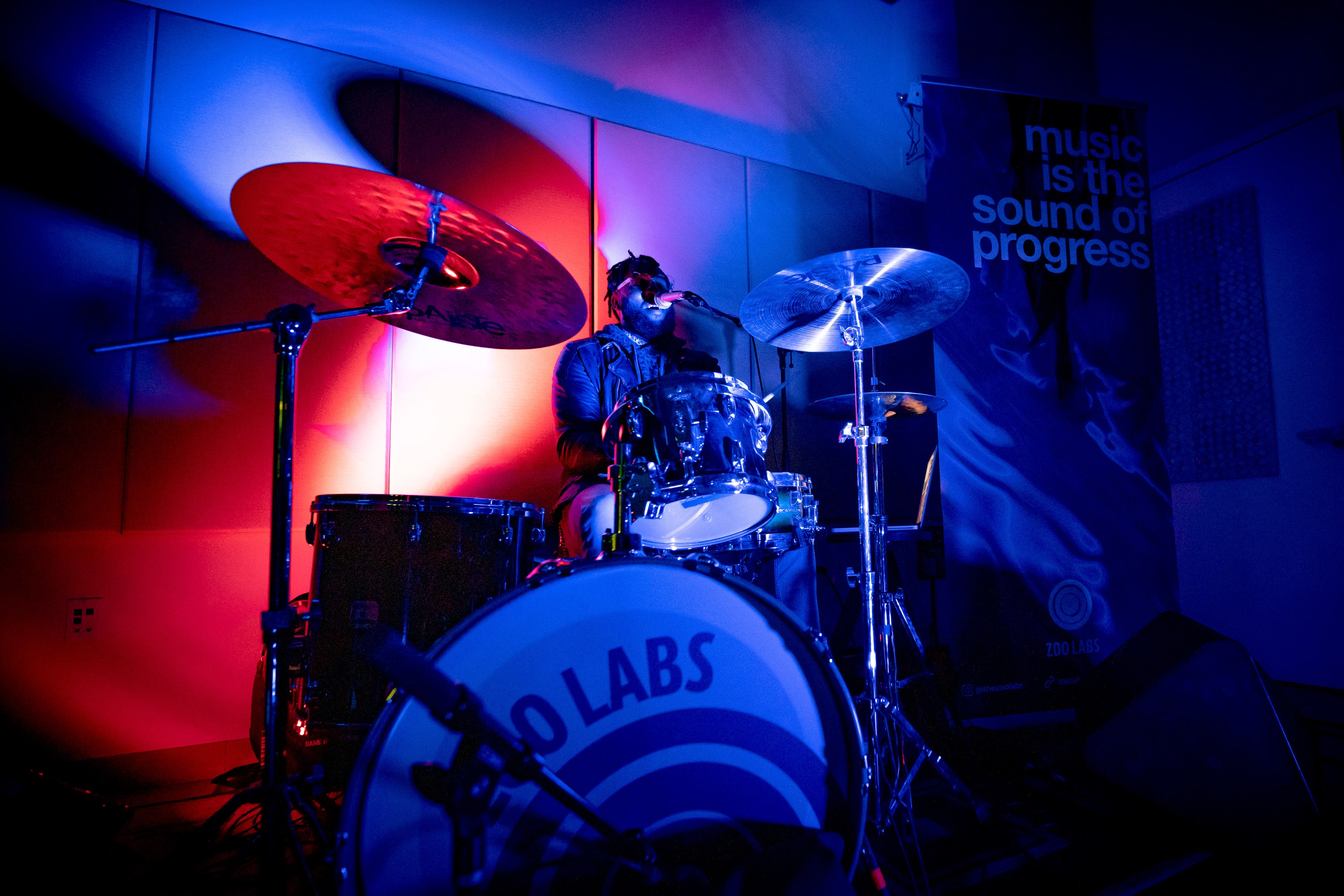 Artist Dame Drummer plays the drums and sings into a microphone in a dark room with moody blue and red lighting