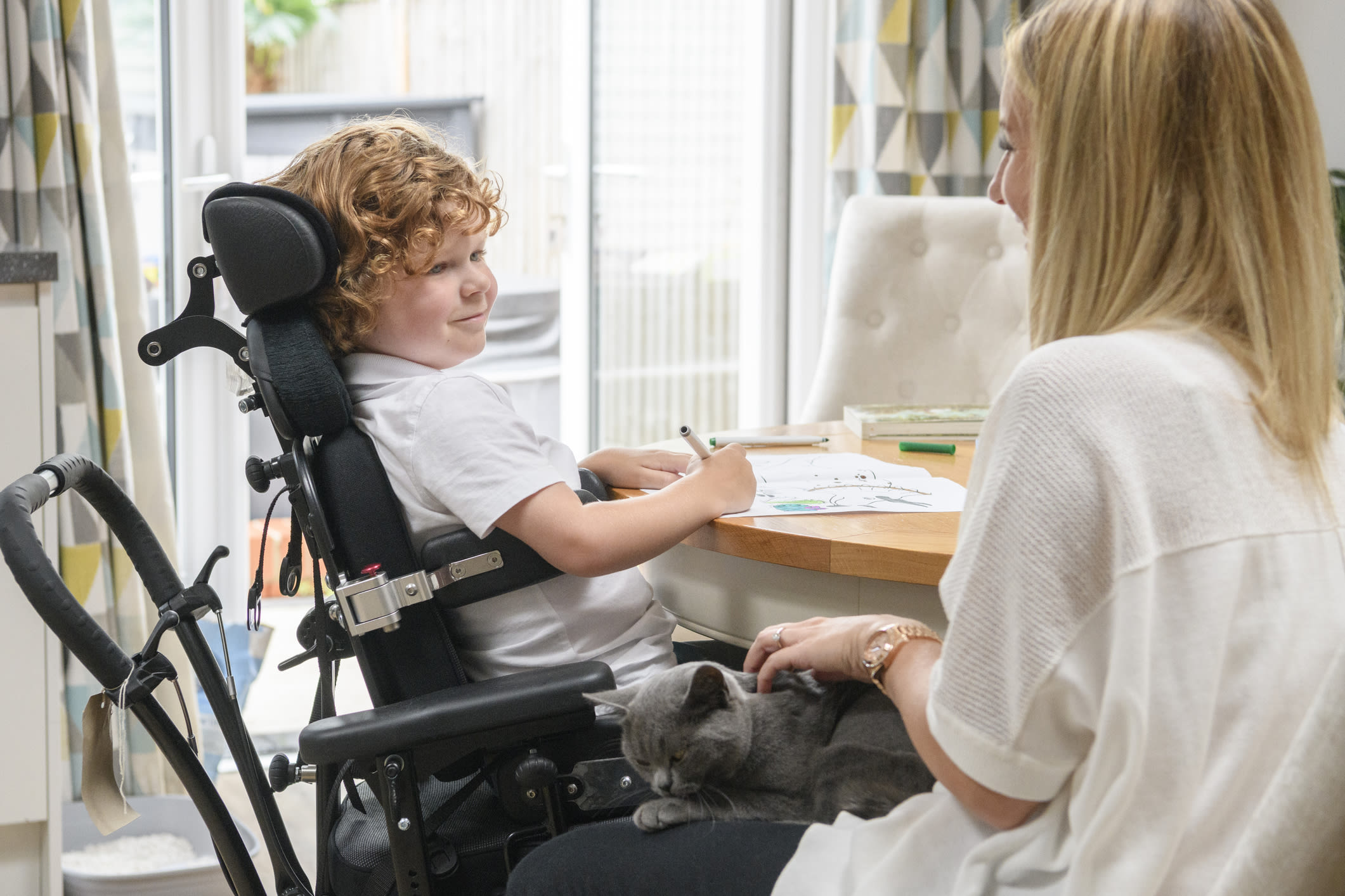 picture of a boy with red curly hair in a white t-shirt sitting at a table with paper & pencil in had.  He uses a wheel chair.  Sitting next to him is a blond haired woman wearing a white shirt with a grey cat sitting on her lap. 