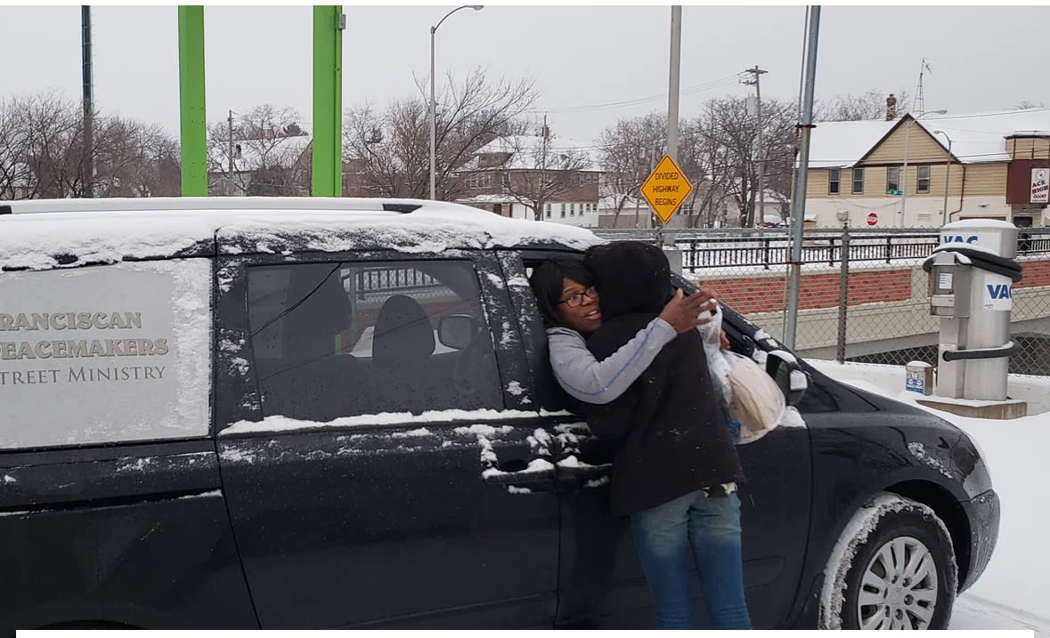 A woman in a van hugging a woman on the street with a lunch in her hand. 