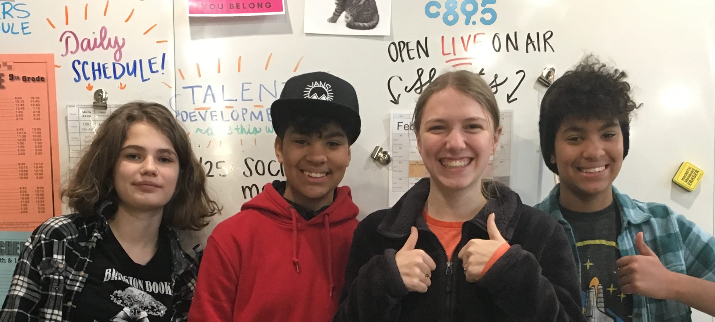 A photo of 4 smiling young people standing in front of a large whiteboard. Two of the students have long, straight hair and pale skin, the other two students are identical twins with brown skin and short, curly hair. One of the students with straight hair is giving the thumbs up sign with both hands and one of the students with curly hair is also giving the thumbs up sign. The whiteboard has the handwritten words "Daily Schedule", "TalentDevelopment", "Open Live On Air", and a c895 bumper sticker.