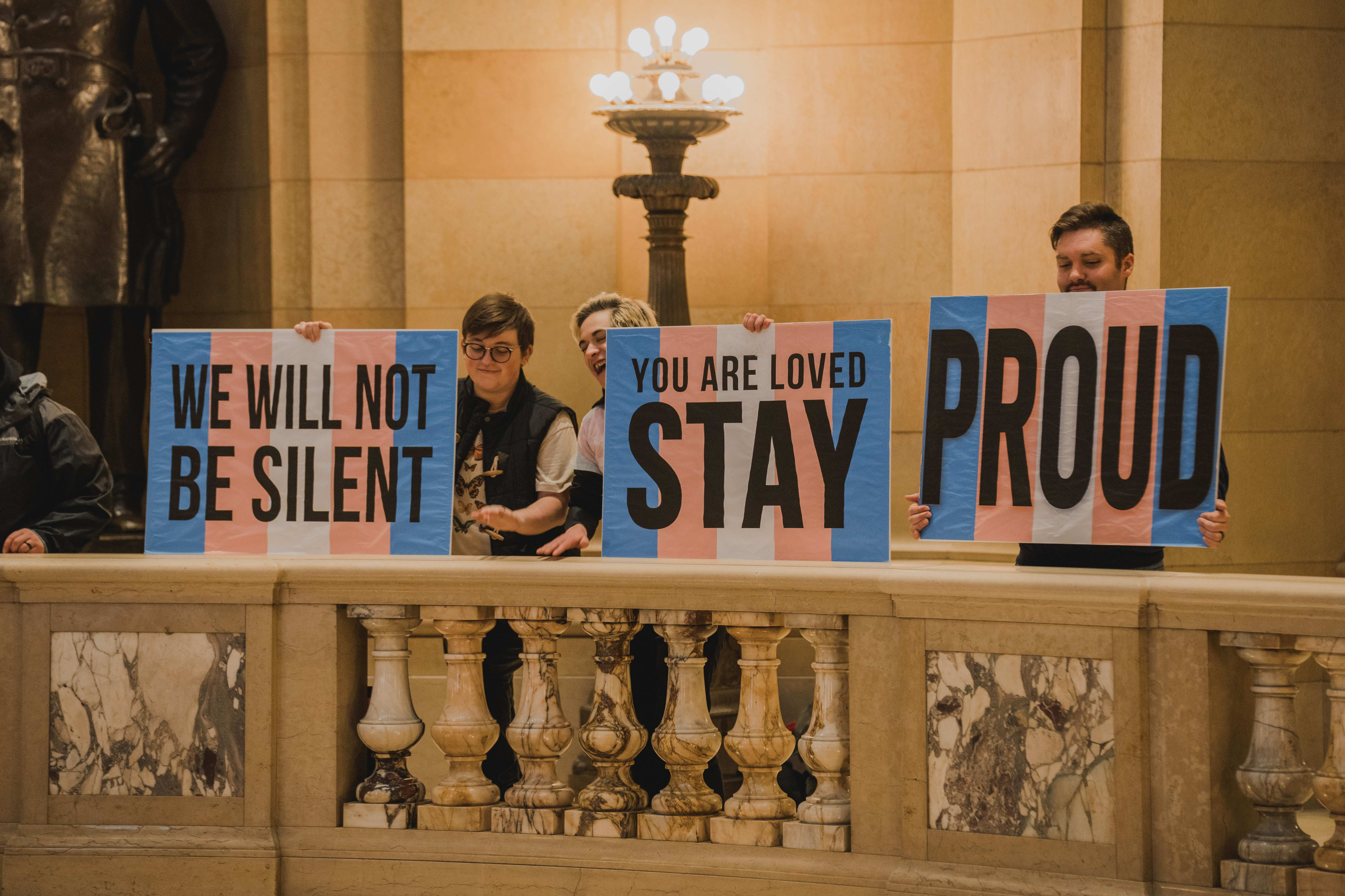 A photo from OutFront Minnesota's Trans Day of Visibility event, with three organizers holding up protest signs with the trans flag colors on them on the balcony of the state capitol. 
