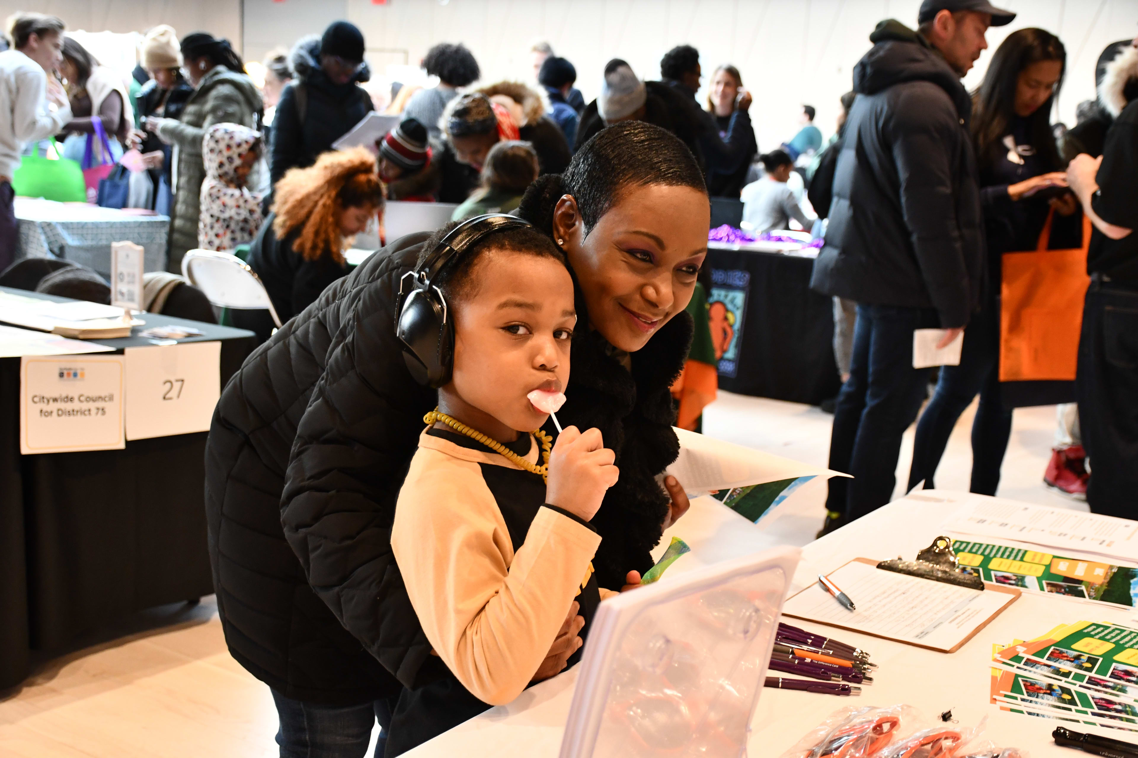Young brown skinned boy wearing headphones looks intently at the camera while enjoying a lollipop, a smiling dark skinned woman leans over next to him at a table with resource materials