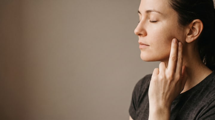 A woman in profile with her eyes closed, resting two fingertips on her jaw below the ear.