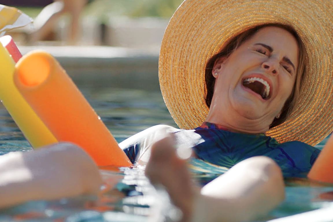 Woman in a wide-brimmed sun hat floating in the pool laughing, surrounded by pool noodles