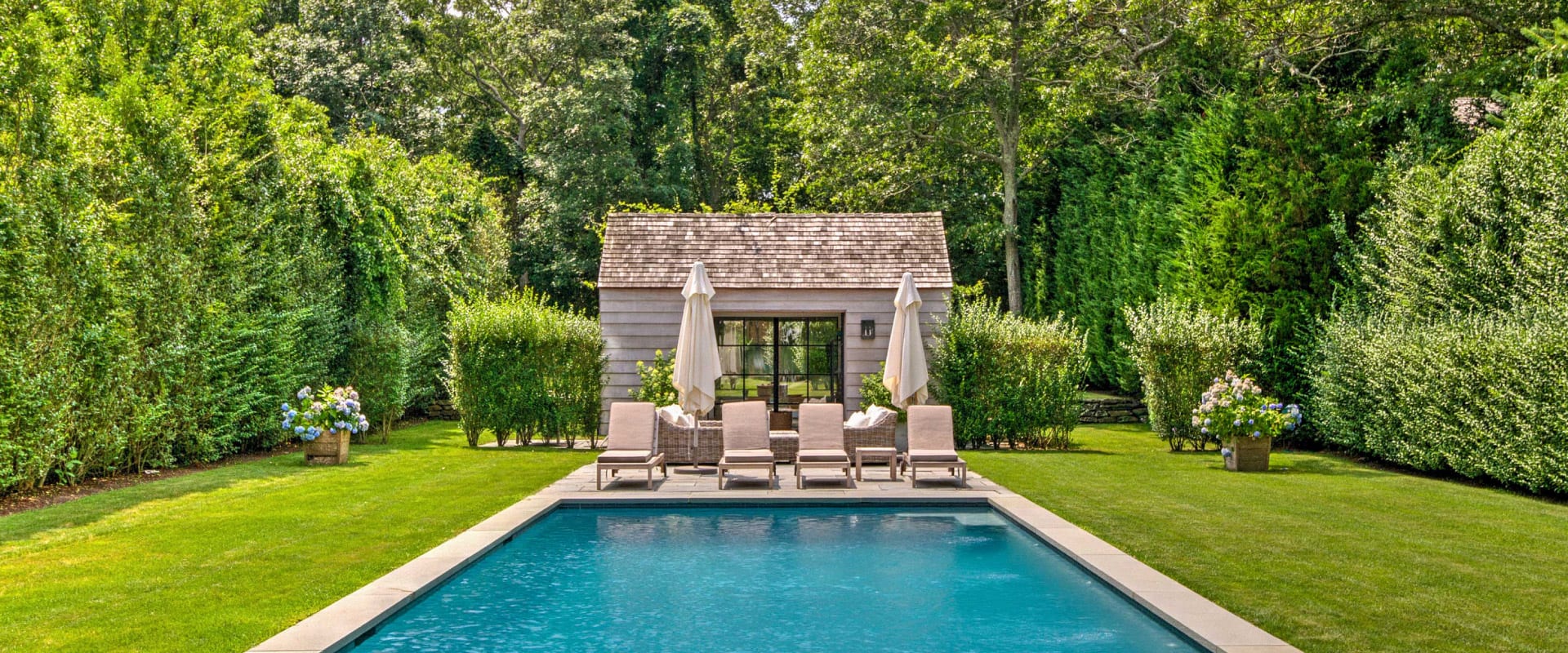 Summer backyard with a pool, pool chairs, and pool house surrounded by bushes and trees, creating a great guest experience with one of the best rental backyard ideas