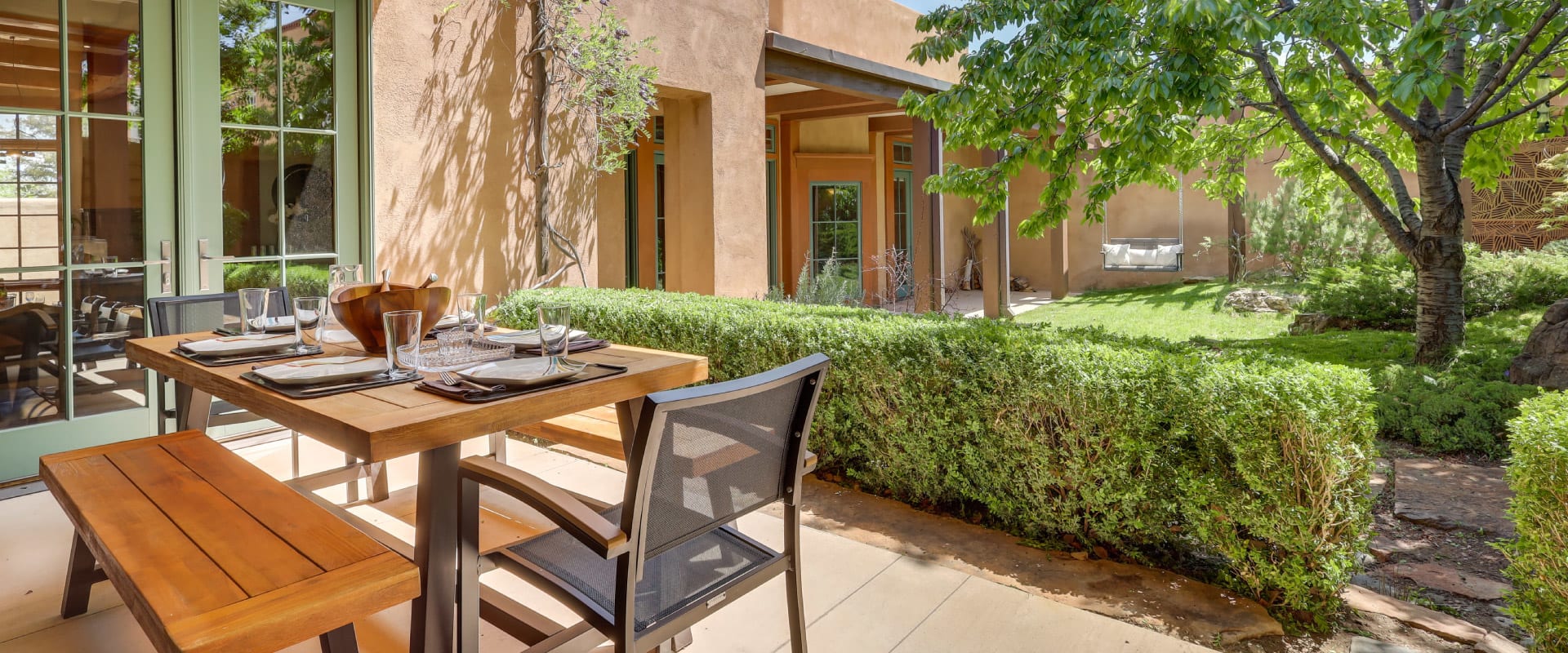 Backyard porch of southwest style home with an outdoor dining table surrounded by bushes and trees.