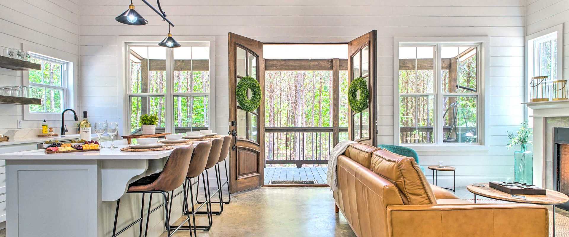 Interior shot of a modern, white, cabin, living room and kitchen with double doors opening to the porch and trees in the backyard.