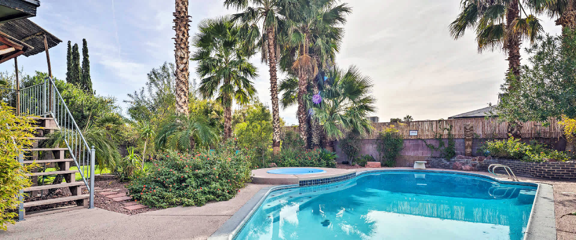 Large outdoor pool and hot tub located in the backyard of a vacation rental property, surrounded by palm trees and flowering bushes.