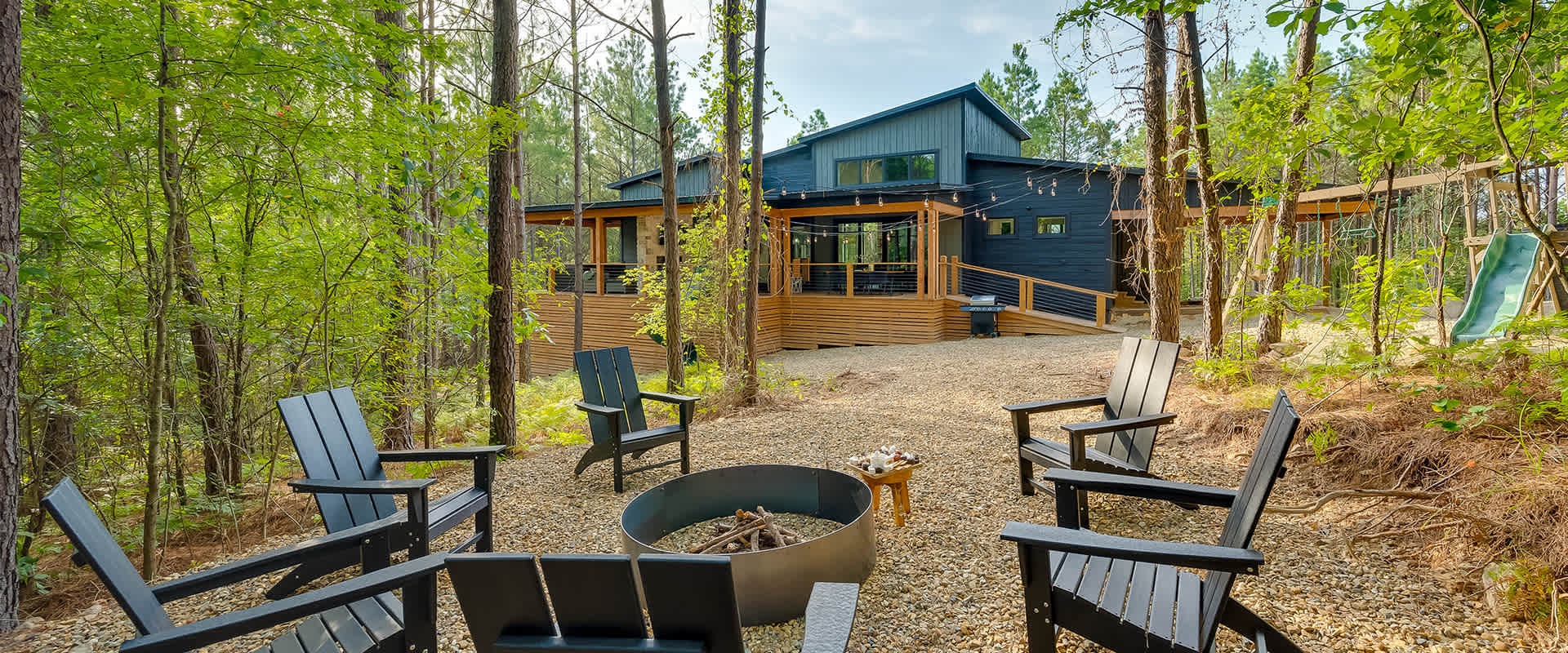 Exterior view of the "Aces Wild" vacation rental in Broken Bow, OK with a firepit surrounded by chairs in the foreground and a slide and swingset amongst the trees