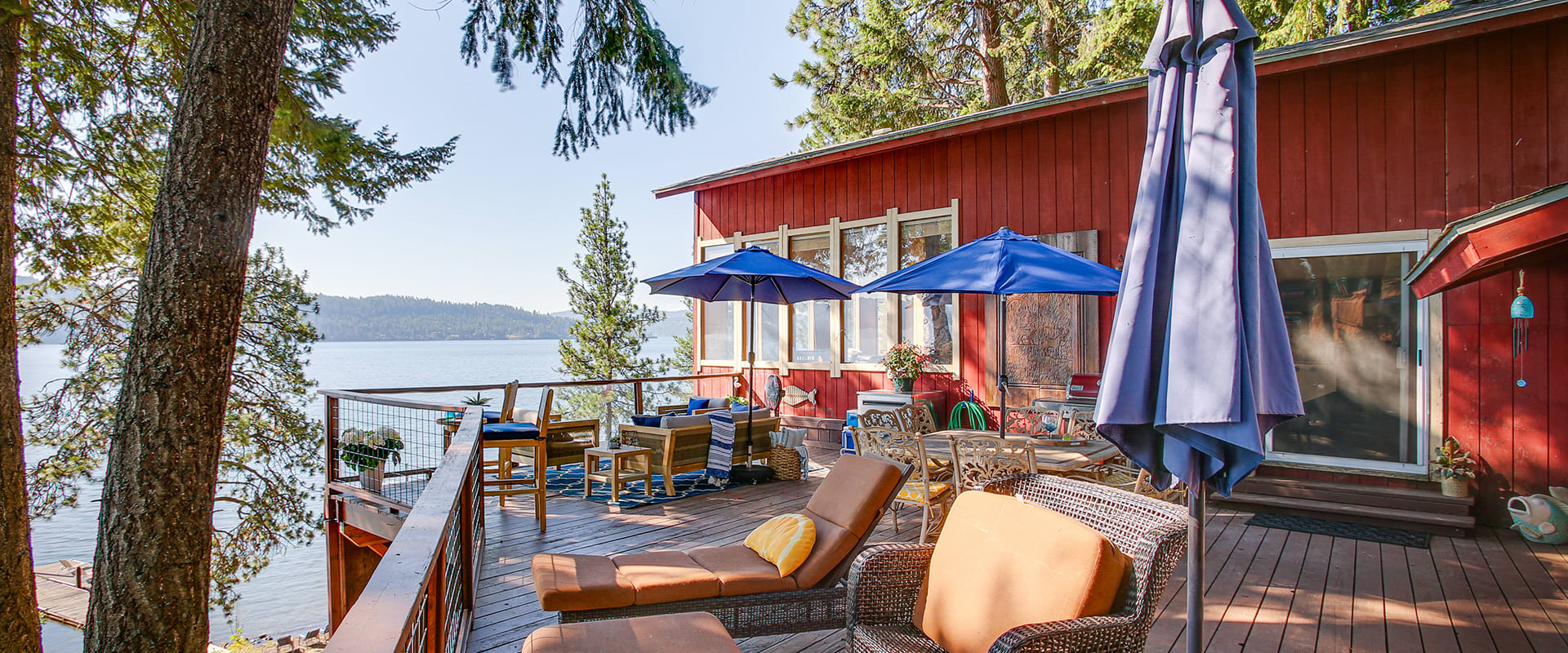 Exterior view of a 3-bedroom, 1-bath cabin in Coeur d'Alene, ID with a spacious deck with multiple tables and umbrellas overlooking a serene lake — the ideal space for morning coffee or evening relaxation.