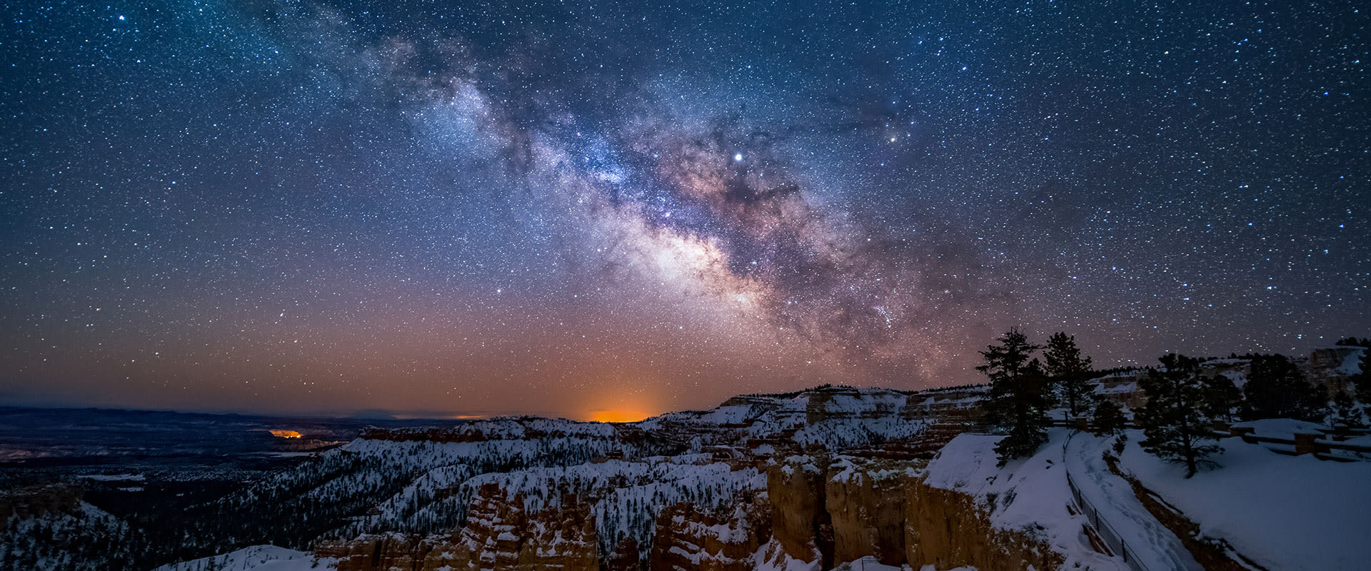 Panoramic photo of Bryce Canyon National Park with a glowing Milky Way in the night sky lighting up freshly fallen snow.