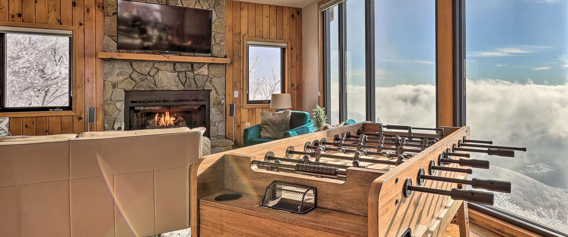 Photo of a cozy mountain cabin living room with a large stone fireplace and a wall-mounted TV. The space includes comfortable seating and a foosball table, along with floor-to-ceiling windows that provide a breathtaking view of snow-covered trees and a sea of clouds.