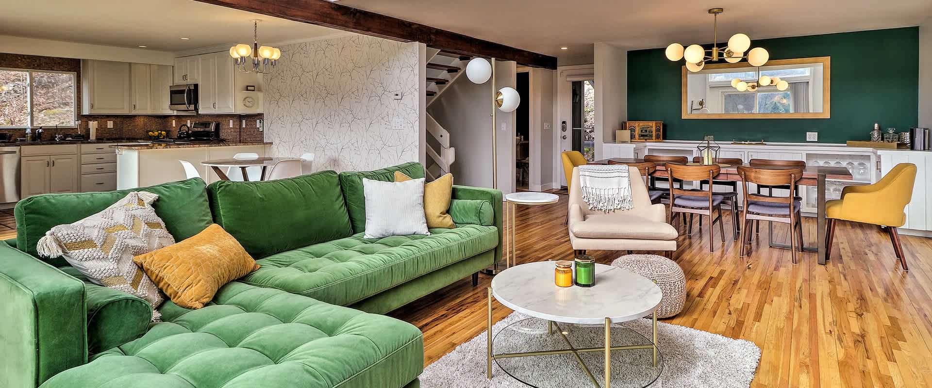Photo of an open-plan living and dining area featuring a vibrant green sectional sofa, beige armchair, round marble coffee table, and a stylish dining table with wooden chairs. The kitchen in the background has white cabinetry and stainless steel appliances.