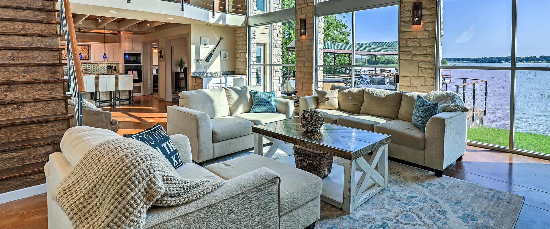 Photo of a spacious and modern living room with large windows overlooking a lake. The room features comfortable beige sofas, and a stylish wooden coffee table, with a staircase leading to the upper floor and a kitchen area with bar seating.