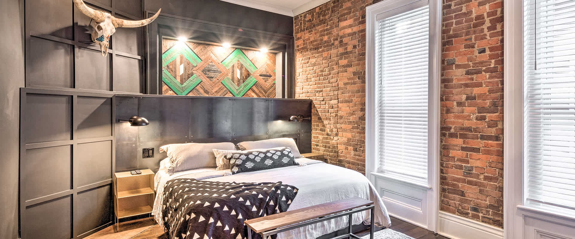 Photo of a bedroom with a large bed against a dark wooden backdrop with a mounted cow skull, all next to an exposed brick wall.