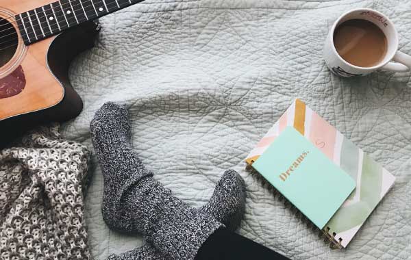 A notebook, coffee cup, and guitar surround a person's feet on a bedspread