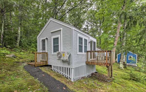 Tiny home with elevated back porch situated on the side of a lush forest mountainscape in New England, with an additional bright blue building in the background