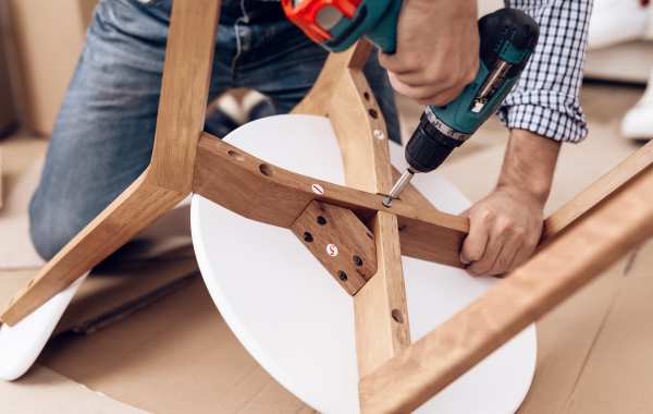 Close up of a man using a drill to tighten the screws on the bottom of a white chair with wooden legs