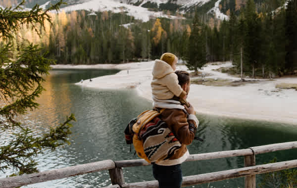 Image of a man hiking with a toddler child on his shoulders. Man and child are dressed warmly, standing on a bridge with snowy mountain and lake scenery