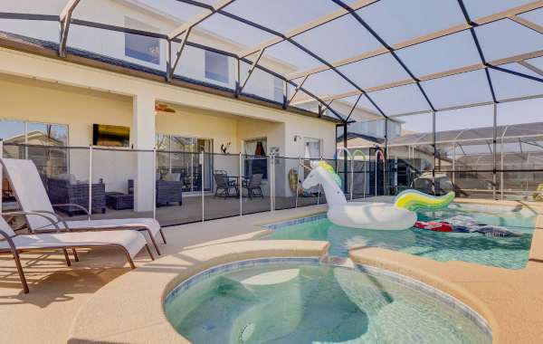 Image of the back exterior of a two-story vacation rental with a covered patio and outdoor furniture, along with a fenced pool and hot tub covered by a domed ceiling. The pool area has two lounge chairs and floaties