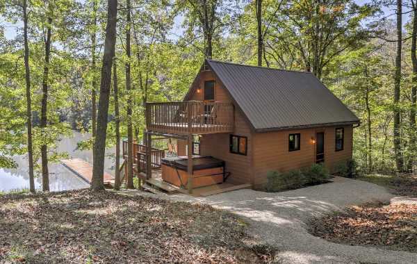 Multi-story cabin surrounded by trees with vacation rental amenities like an outdoor hot tub and boat dock leading out to a lake