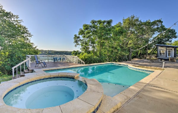 Exterior view of a tiny home with a private pool, hot tub, and small deck surrounded by green trees and a blue sky.