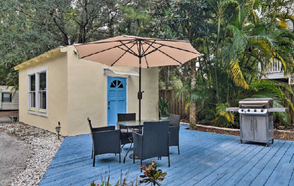 Exterior view of a yellow stucco tiny home with a outdoor table and chairs, umbrella, and grill on a blue deck surrounded by palm trees.