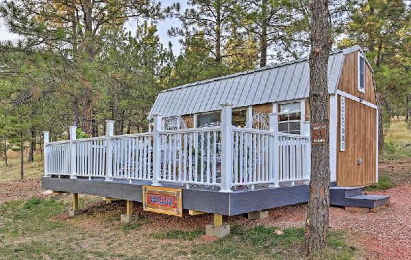 Exterior view of a barn-style tiny home with a white picket fenced porch and Western signage, surrounded by a pine tree forest.