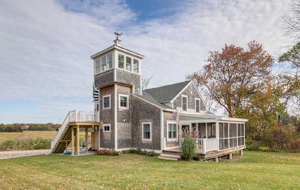 A vacation rental home in Scituate, MA with a tower and screened porch sits in a grassy yard surrounded by trees under a partly cloudy sky.