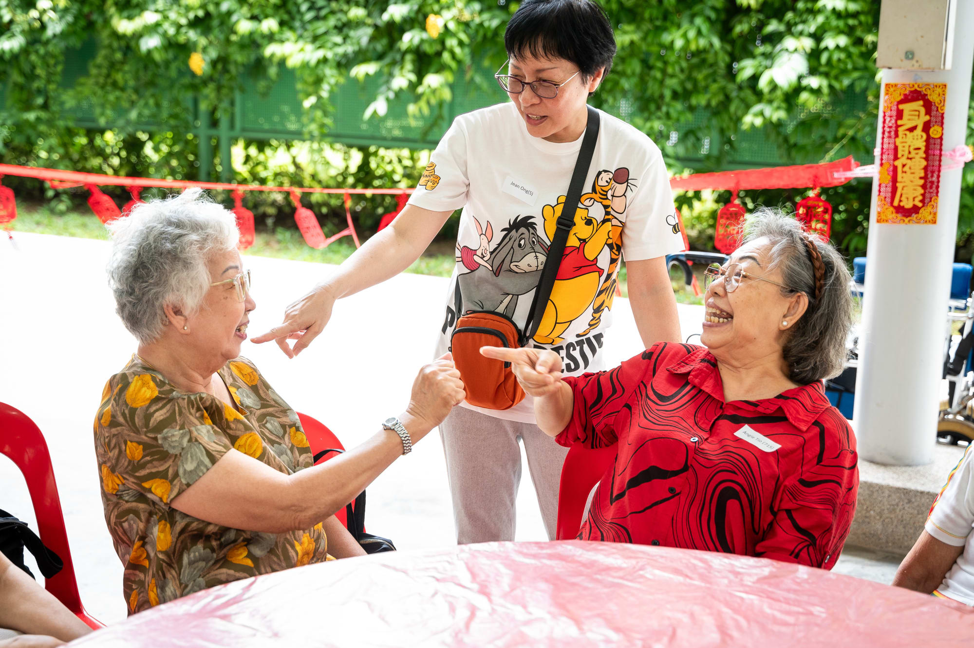 Two seniors laugh and point at each other across a table while a volunteer leans in to share the joke