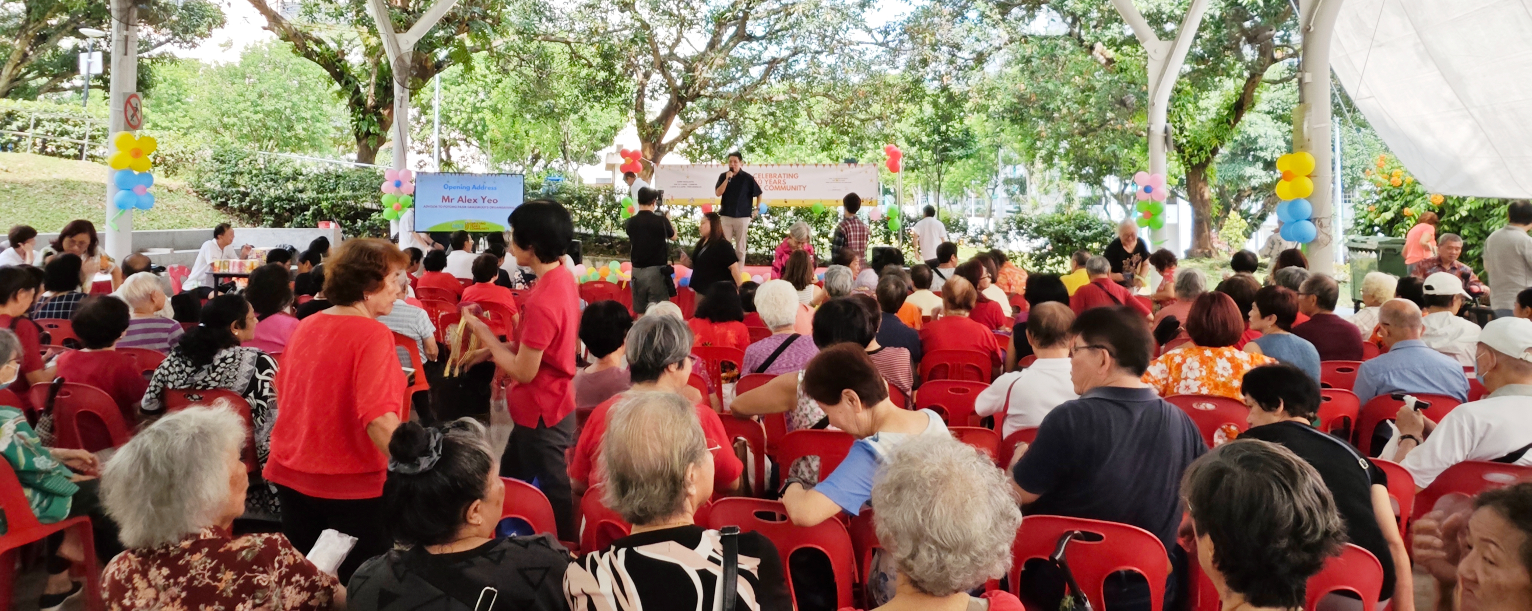A community event with people seated on red chairs under a canopy. A speaker addresses the crowd. Colorful balloons decorate the area.